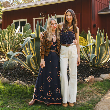 Two women stand side by side outdoors in front of large succulents and a red building, wearing bohemian-style outfits, one in a patterned dress and the other in a sleeveless top and white pants.