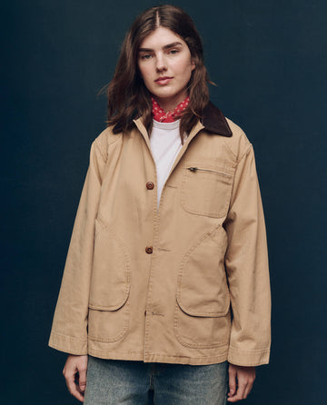 Amidst a dark backdrop, a person exudes outdoor fashion elegance in THE GREAT. SP25 COLLECTORS' The Barn Jacket - Golden, featuring a distinguished brown collar and accessorized with a chic red scarf.