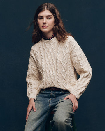 A person with long hair wearing The Classic Cable Pullover in cream from THE GREAT. SP25 COLLECTORS and jeans sits against a dark background.