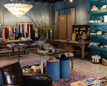 A stylish boutique interior featuring clothing racks, a large chandelier, green tiled walls, wooden tables with various products, and decorative elements on a patterned rug.