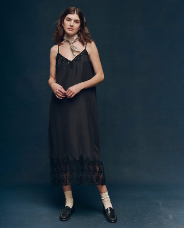 A woman stands against a dark background wearing The Legacy Dress - Black by THE GREAT. SP25 COLLECTORS, showcasing elegant fashion elements like a scarf around her neck, yellow socks, and black loafers.