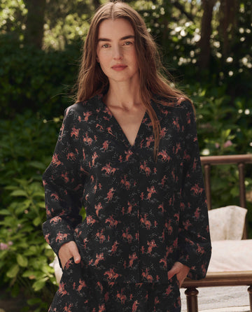 A woman with long brown hair stands outdoors by green foliage and a wooden chair, wearing THE GREAT. SP25 SLEEP's The Pajama Shirt in Navy Cowboy.