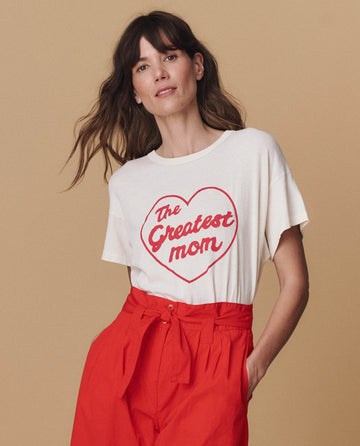 A woman poses against a beige background wearing THE GREAT. SP25 MOM GRAPHICS The Boxy Crew in washed white with The Greatest Mom graphic and high-waisted red pants—showcasing the perfect mom graphic look.