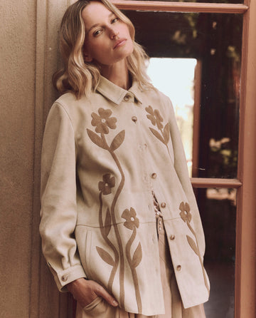 Someone wearing THE GREAT. SP25 D1's The Floral Applique Suede Jacket in Tonal Desert Tan stands by a wooden door with glass panels, gazing at the camera.