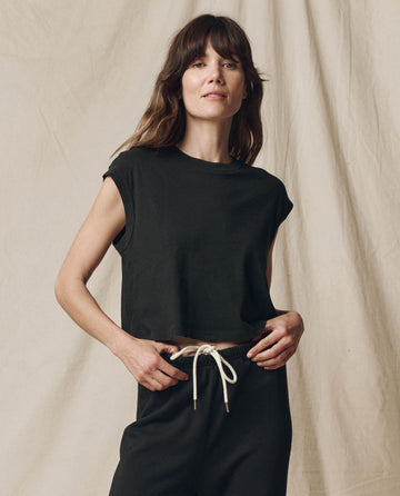 A person with long hair wearing The Square Tee in Almost Black, by THE GREAT. SU23 CORE KNITS, and black pants with a white drawstring, reminiscent of vintage athletic style, stands against a plain beige backdrop.