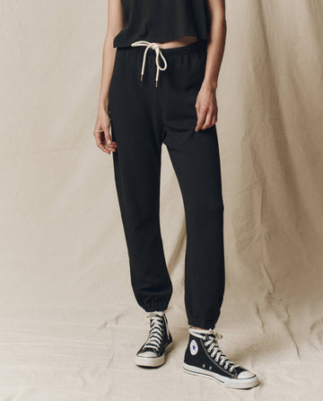 A person stands against a beige background wearing The Stadium Sweatpant by THE GREAT. CORE KNITS in solid Almost Black, paired with high-top sneakers and featuring an elasticated waist.