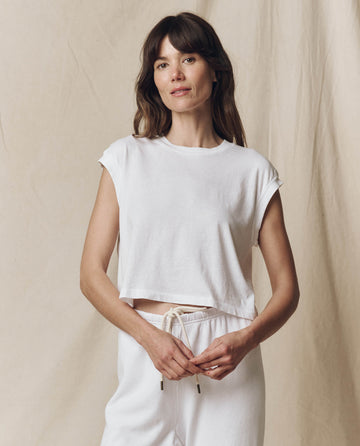 A person with long hair stands against a plain background, confidently wearing THE GREAT. SU23 CORE KNITS’ The Square Tee in True White along with white pants, looking at the camera.