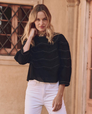 A person with long blonde hair in The Beaded Chevron Top by THE GREAT. SP25 D1 and white pants stands in front of a building with lattice windows, embodying the essence of women's fashion.