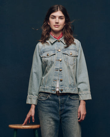Wearing THE GREAT. SP25 COLLECTORS' The Slouchy Jean Jacket in Great Plains Wash, a person embodies a vintage-inspired look with matching jeans, standing beside a stool against a dark background.
