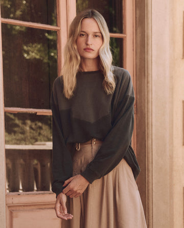 A person with long blonde hair slouches in front of a window, wearing THE GREAT. SP25 D1's The Western Slouch Sweatshirt in Washed Black and a beige skirt. They look directly at the camera, exuding a relaxed western charm.