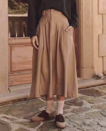 A person stands outside in The Western Field Skirt - Barley by THE GREAT. SP25 D1, paired with a black sweater, maroon shoes, and white socks. Hands are tucked into the skirt's pockets.