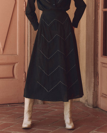 A stylish individual wears THE GREAT.'s black Beaded Chevron Skirt and white cowboy boots, with beaded accessories, standing on a red-tiled floor before wooden doors.