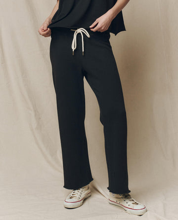 A person in The Wide Leg Cropped Sweatpant in Almost Black by THE GREAT. CORE KNITS stands against a beige background, lightly gripping the waistband of their pants, and wearing white sneakers.