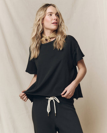 A woman in THE GREAT. CORE KNITS' "The Crop Tee. Solid -- Almost Black" paired with high-rise jeans featuring a white drawstring, and accessorized with a yellow neck scarf stands against a plain backdrop.