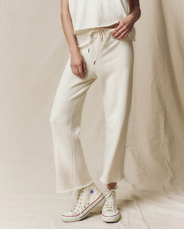 Someone wearing The Wide Leg Cropped Sweatpant in Washed White by THE GREAT. CORE KNITS and a sleeveless top, paired with white high-top sneakers, stands on a neutral fabric backdrop.