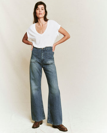 A person in a casual outfit stands against a plain background, wearing a white V-neck T-shirt, The Dock Jean in Marina Wash by THE GREAT. PF24, and brown shoes.