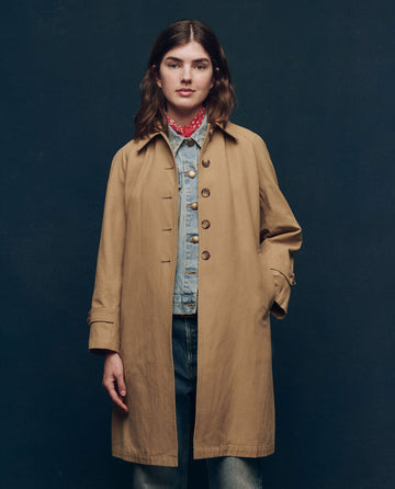 A person presents a chic look by layering The Trench Coat in Khaki from THE GREAT. SP25 COLLECTORS over a denim jacket, with a red bandana around their neck, set against a dark background.