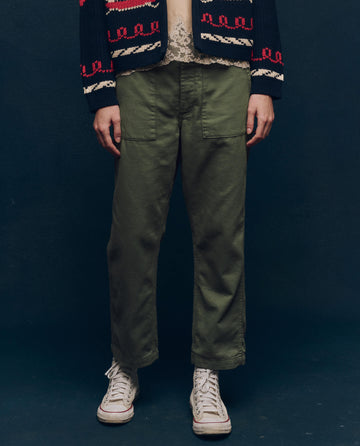 A person wearing The Vintage Army Pant by THE GREAT. SP25 COLLECTORS, a dark patterned jacket, and white high-top sneakers stands against a dark backdrop, exuding a vintage vibe.