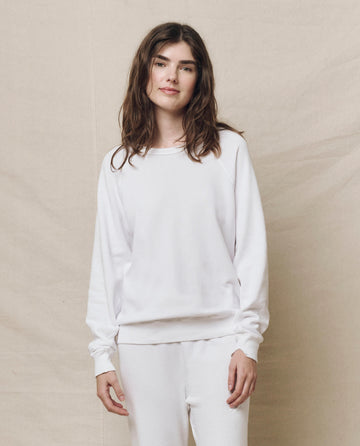 A woman with long hair stands against a plain backdrop, wearing The College Sweatshirt in True White and matching pants by THE GREAT. SP25 KNITS, crafted from double-washed terry.