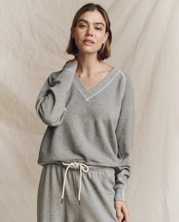 A person wearing The V-Neck Sweatshirt in Varsity Grey by THE GREAT. FALL 24 KNITS and drawstring pants stands against a beige background.