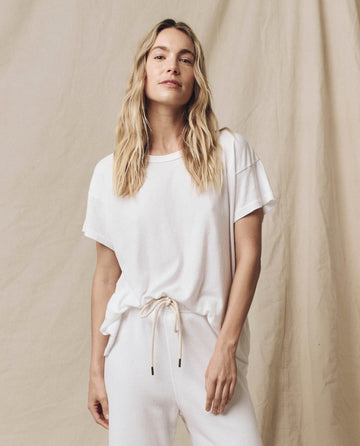 A person with long blonde hair wearing The Boxy Crew in True White by THE GREAT. CORE KNITS and white pants stands in front of a beige backdrop.