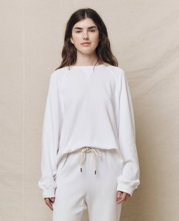 A person stands against a beige background wearing The Slouch Sweatshirt in True White by THE GREAT. HOL 24 KNITS, paired with matching pants, giving off a handcrafted vintage vibe, with their hands casually by their sides.