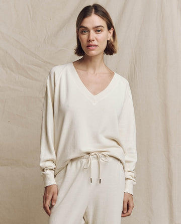 A person with short hair is standing against a beige fabric background, wearing THE GREAT. FALL 24 KNITS' "The V-Neck Sweatshirt" in washed white and drawstring pants.