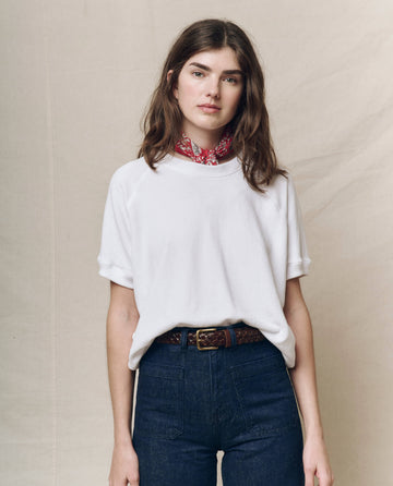 A woman in THE GREAT. SP25 KNITS' Short Sleeve Sweatshirt in True White, blue jeans, and a red patterned scarf stands against a neutral background. Her hands rest in her pockets, highlighting this perfect spring transition piece that combines comfort and style.