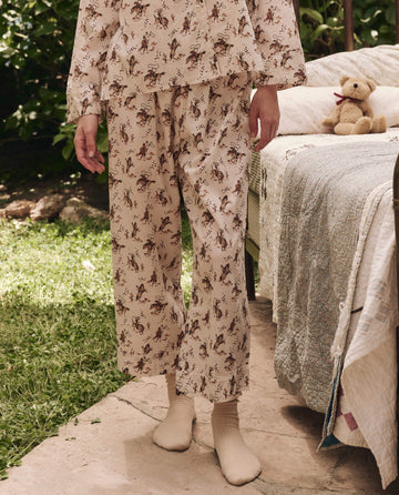 Standing outdoors on a stone path, a person wears THE GREAT. SP25 SLEEP's Easy Sleep Pant in Cream Cowboy—comfortable, cream-colored pajamas with a horse print—paired with light socks. A quilted bed and teddy bear create cozy vibes.