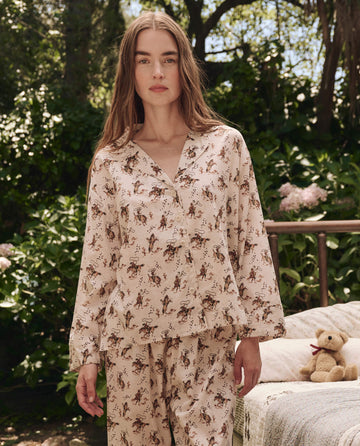 A woman stands outdoors wearing THE GREAT. SP25 SLEEP's The Pajama Shirt in Cream Cowboy, featuring a brown animal print; behind her, a teddy bear rests on a bench surrounded by greenery.