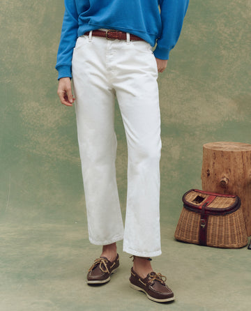 A person wearing The Wayne Jean in True White by THE GREAT. SP25 D1, a blue sweatshirt, and brown boat shoes stands indoors beside a wicker picnic basket and wooden stump with a green textured backdrop.