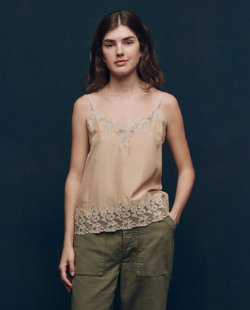 Against a dark background, a woman stands wearing "The Legacy Top” by THE GREAT. SP25 COLLECTORS in nude, featuring a stylish lace-trimmed camisole design, paired with green pants.