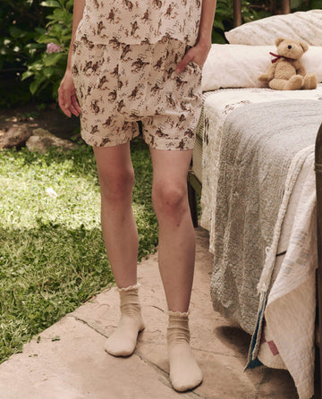 A person stands on a stone path in an outdoor garden square, wearing THE GREAT. SP25 SLEEP's The Square Pajama Short in Cream Cowboy, with a matching top and socks, beside a bed topped with a quilt and teddy bear.
