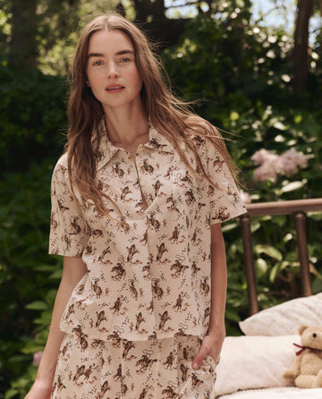 A woman wears THE GREAT. SP25 SLEEP's The Short Sleeve Pajama Shirt in Cream Cowboy, paired with matching shorts, standing outdoors by a bed adorned with pillows and a teddy bear amid lush greenery.