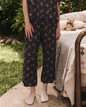 Outdoors, a person stands by a bed wearing THE GREAT. SP25 SLEEP The Easy Sleep Pant in Navy Cowboy and light socks; a teddy bear rests on the bed.