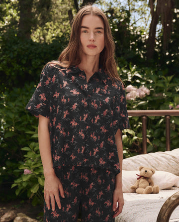 A woman stands outdoors wearing THE GREAT. SP25 SLEEP The Short Sleeve Pajama Shirt in Navy Cowboy; a teddy bear sits on a bench in the background.