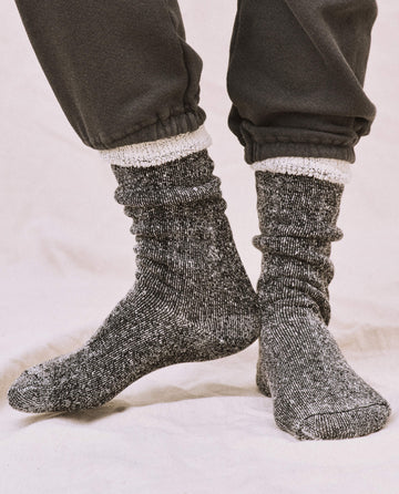 A person stands on a light-colored surface wearing black THE GREAT. SP25 SWEATSHIRT SOCKS, a cozy sweatshirt, and dark pants with elastic cuffs.