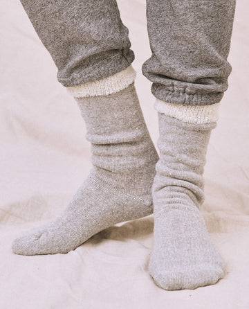 A person stands on a soft beige surface wearing THE GREAT. SP25 SWEATSHIRT SOCKS in Heather Grey, paired with a cozy gray sweatshirt and matching sweatpants.
