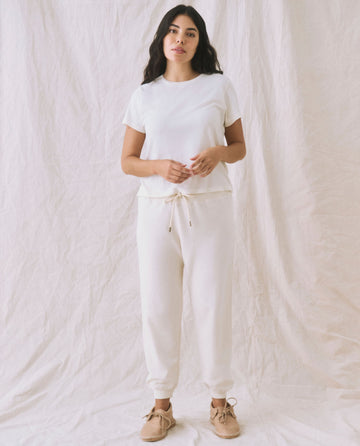 A person stands against a plain background, wearing a white short-sleeve shirt and The Cropped Sweatpant in Washed White by THE GREAT. CORE KNITS, complemented by light brown shoes.