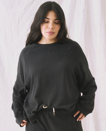 A woman with long dark hair, dressed in The Slouch Sweatshirt by THE GREAT. CORE KNITS in an almost black shade, stands against a light background, exhibiting an extra slouch paired with pants.
