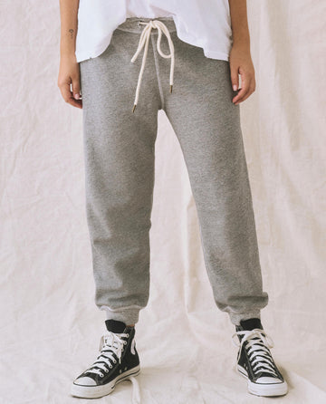 A person standing against a plain backdrop wears The Cropped Sweatpant. Solid by THE GREAT. CORE KNITS in Varsity Grey, featuring ribbed cuffs and a drawstring waistband. They complete the look with a white top and black high-top sneakers. The person's face is not visible.