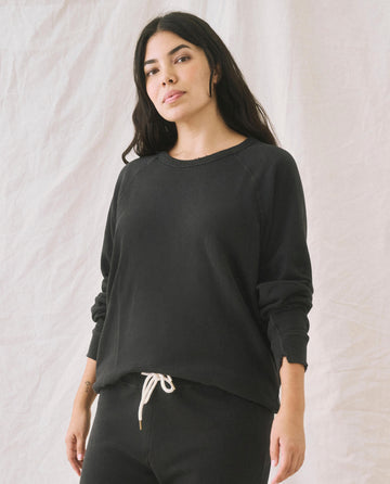 A person wearing The College Sweatshirt in "Almost Black" by THE GREAT. CORE KNITS and black pants with a white drawstring stands against a light-colored fabric backdrop, giving off an oversized sweatshirt vibe.