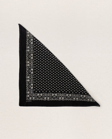 Displayed on a plain light background, THE GREAT. SP24 D2's product, The Bandana in small, features an ultra-soft tri-folded fabric made from 100% cotton in an almost black shade with intricate white floral and star patterns adorning the edges and center.