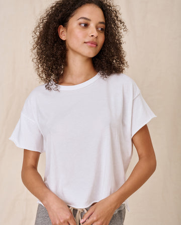 A person with curly hair, wearing The Crop Tee in True White from THE GREAT. CORE KNITS, and high-rise jeans looks to the side against a plain background.