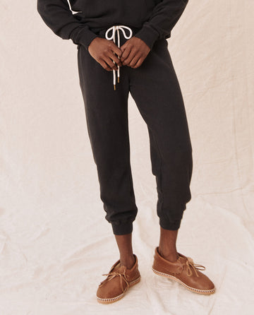 A person in "The Cropped Sweatpant. Solid -- Almost Black" from THE GREAT. CORE KNITS, paired with a dark hoodie and vintage gym pants with ribbed cuffs, stands on a light fabric surface with hands clasped and wearing brown moccasin-style shoes.