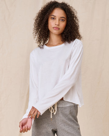 Person with curly hair, wearing The Long Sleeve Crop Tee in TRUE WHITE by THE GREAT. CORE KNITS, paired with gray pants, stands against a neutral background.