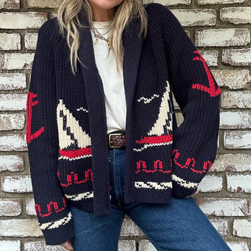 A person wearing a navy blue knit cardigan with red and cream sailboat designs, a white shirt, and blue jeans stands in front of a brick wall.