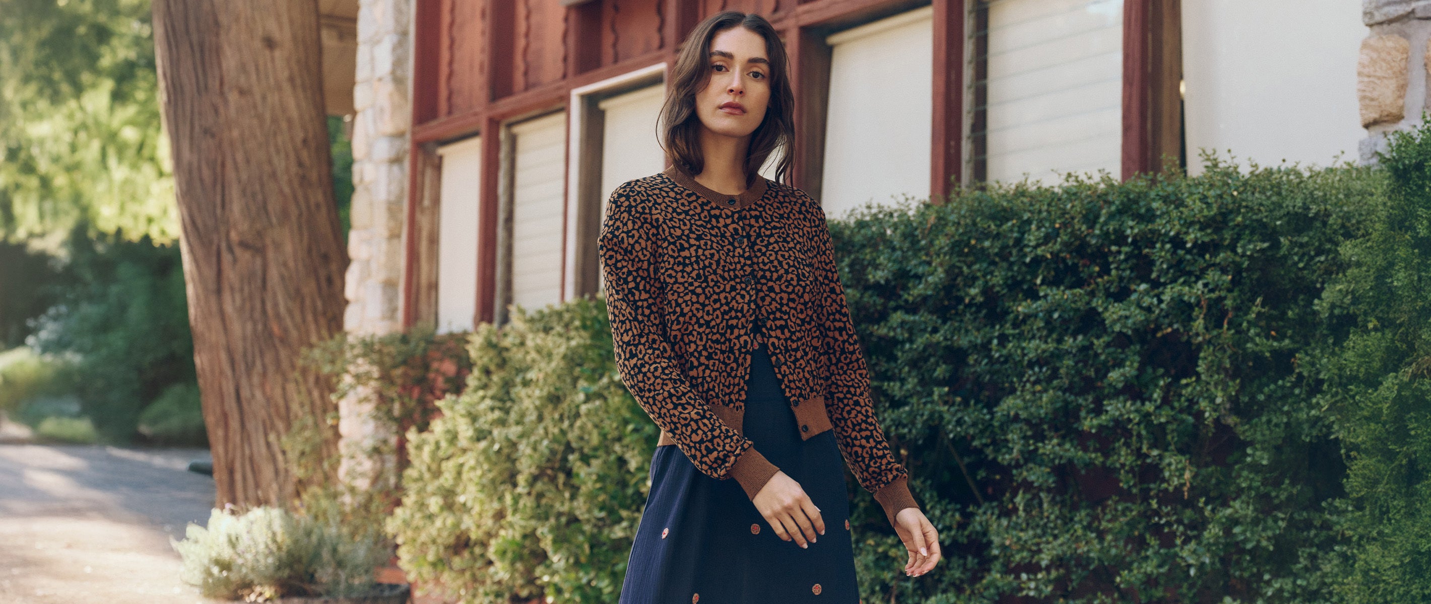 A woman stands outdoors in front of a building with large windows and green shrubs, wearing a patterned long-sleeve top and a dark skirt.