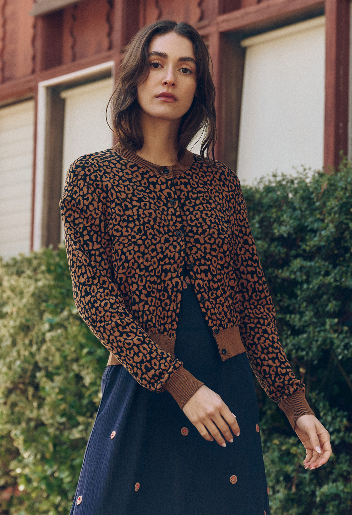 A woman stands outdoors wearing a brown leopard print cardigan and a navy blue dress with small patterns, with greenery and a building in the background.