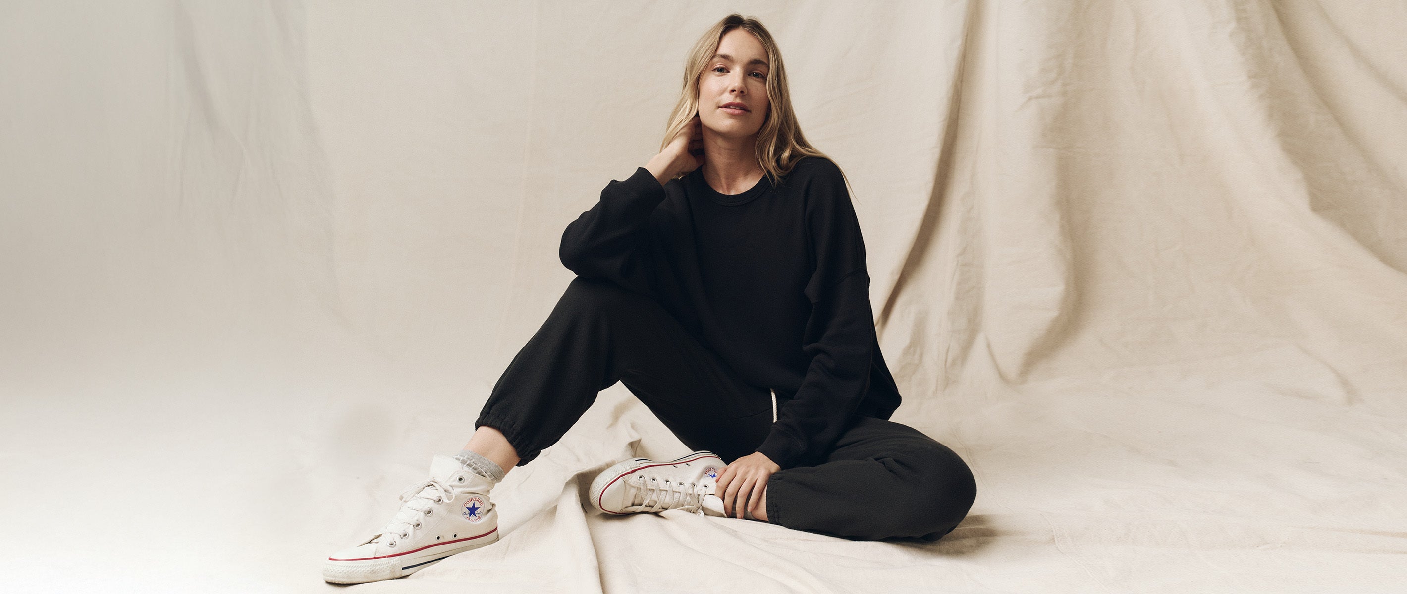 Person sitting on fabric backdrop, wearing a black outfit and white high-top sneakers, with a relaxed pose.